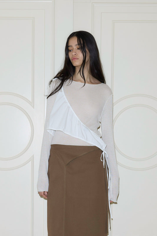 A woman with long dark hair stands before white paneled doors, wearing a sheer beige long-sleeve top, brown skirt, and a white ruffled Cotton Sash draped asymmetrically across her chest and tied at her side.