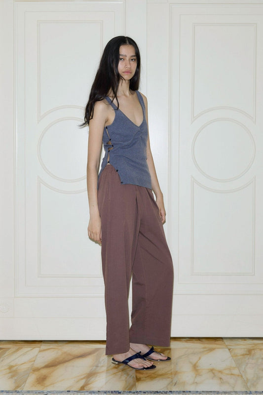 A woman with long dark hair stands on a marble floor, wearing the Ease Trouser in brown, a blue sleeveless top, and black sandals. She poses indoors by white paneled doors, looking slightly toward the camera.