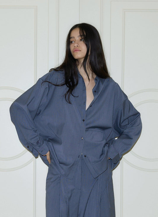 A woman with long dark hair wears the Pocket Wool Shirt in Navy Stripe, styled oversized, paired with matching pants as she stands hands on hips before white paneled doors.