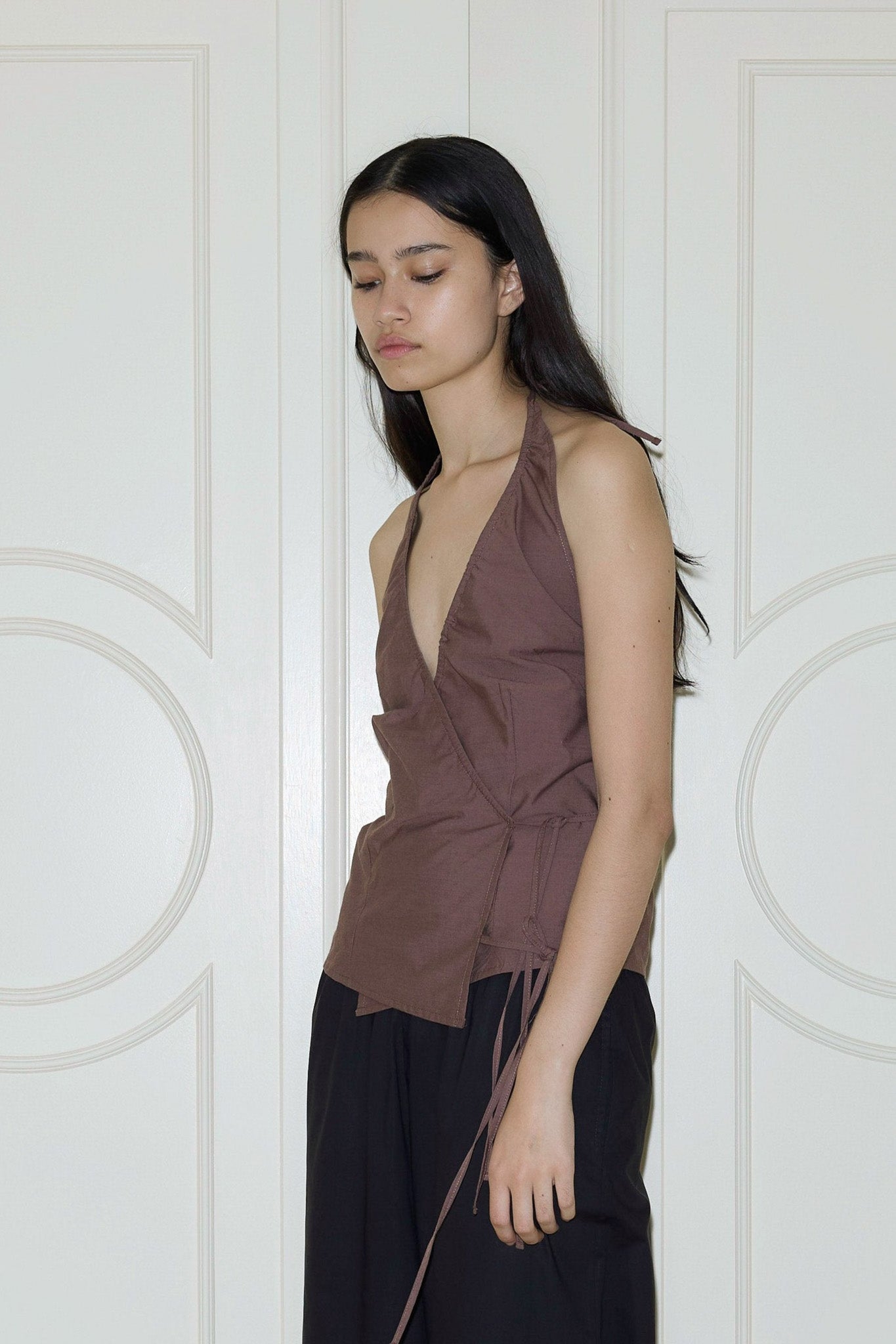 A young woman with long dark hair stands against a cream paneled door, wearing a brown Drawcord Halter and loose black pants, looking down with a neutral expression.