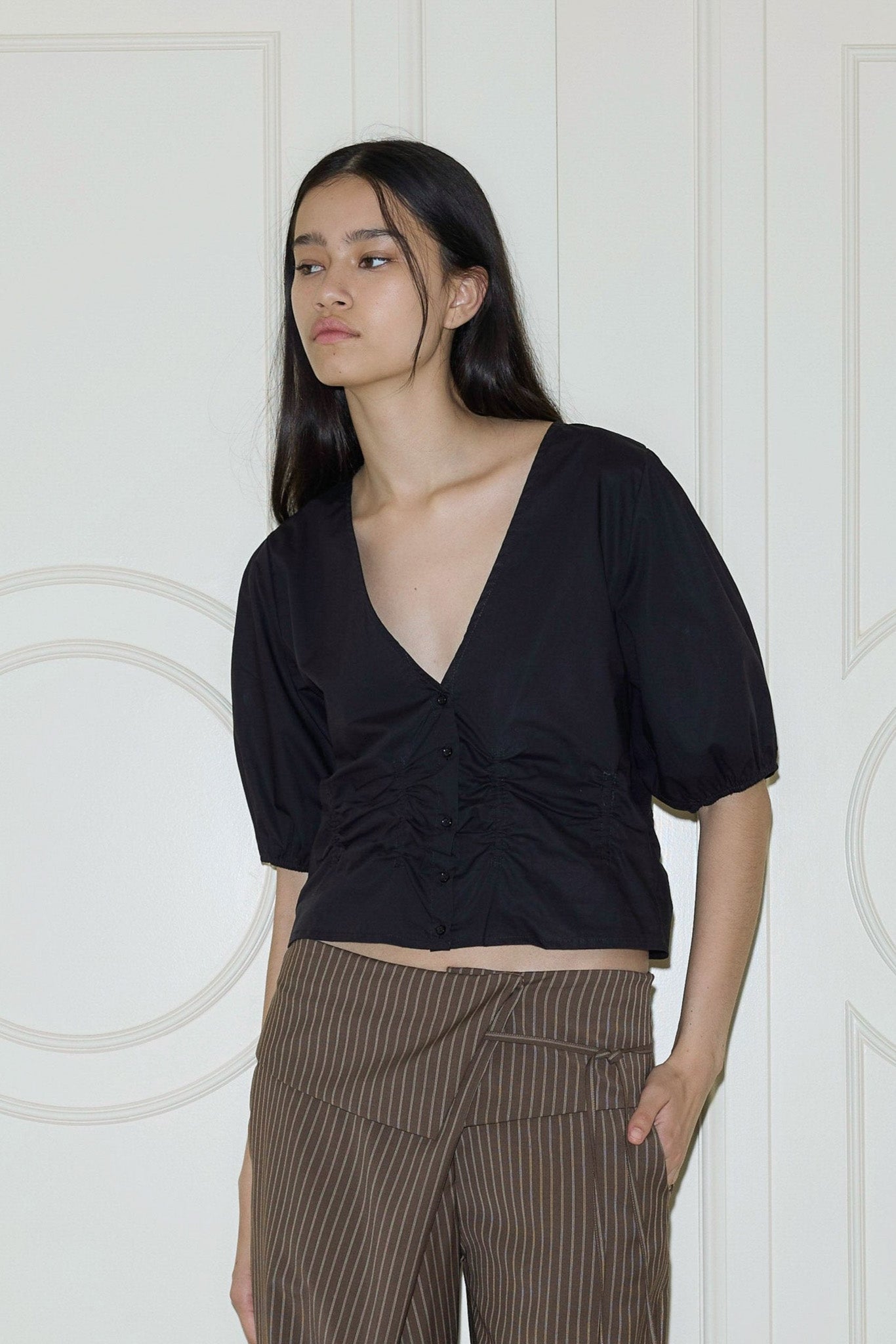 A person with long dark hair wears the Draw Top, a deep V-neck black blouse with short sleeves, paired with brown pinstriped pants, standing by a cream paneled wall.