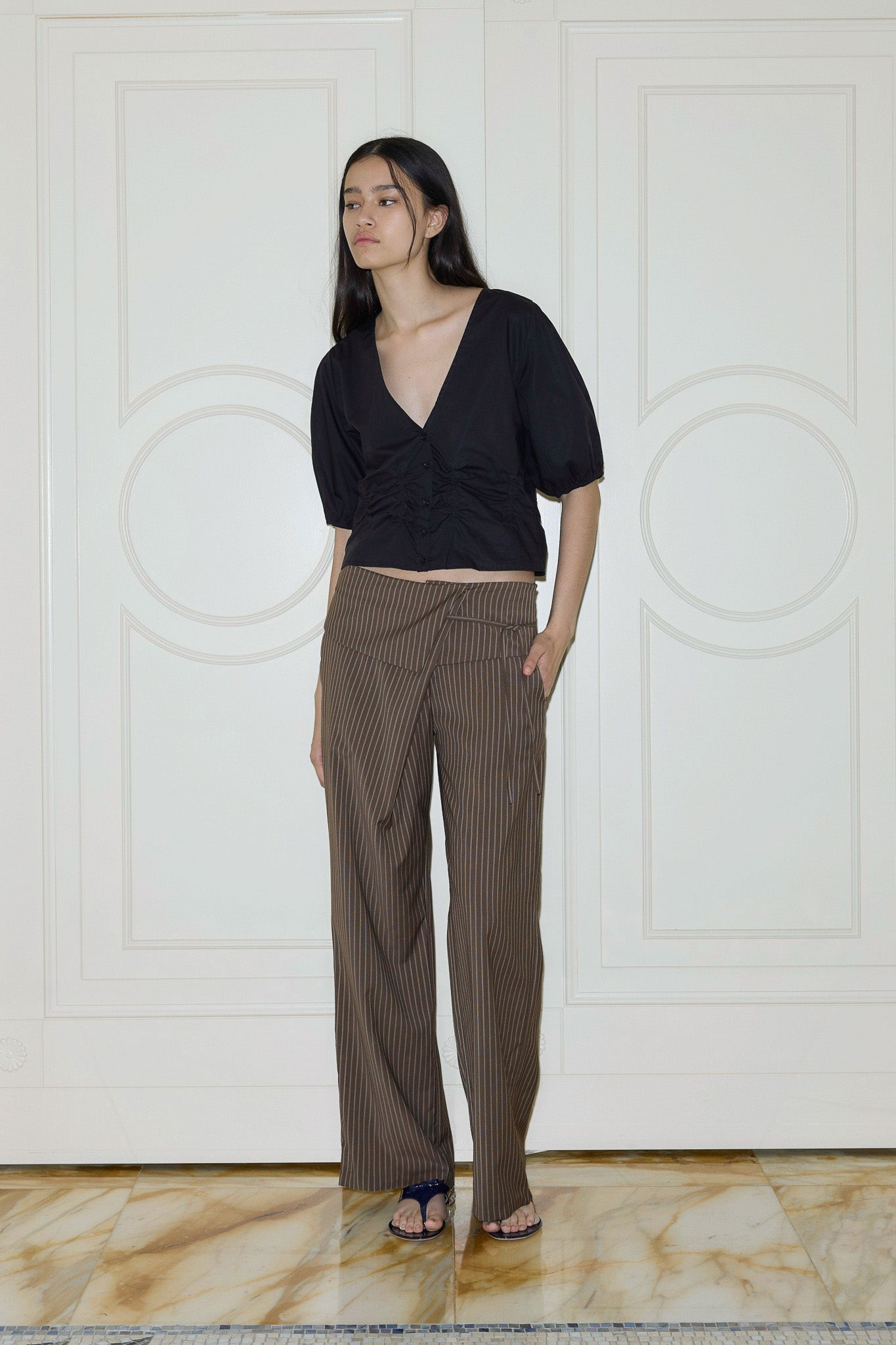 A woman with long dark hair wears a black V-neck blouse, the 0 Folded Wool Pant - Sable Stripe, and black sandals as she stands indoors on a marble floor before white panel doors.