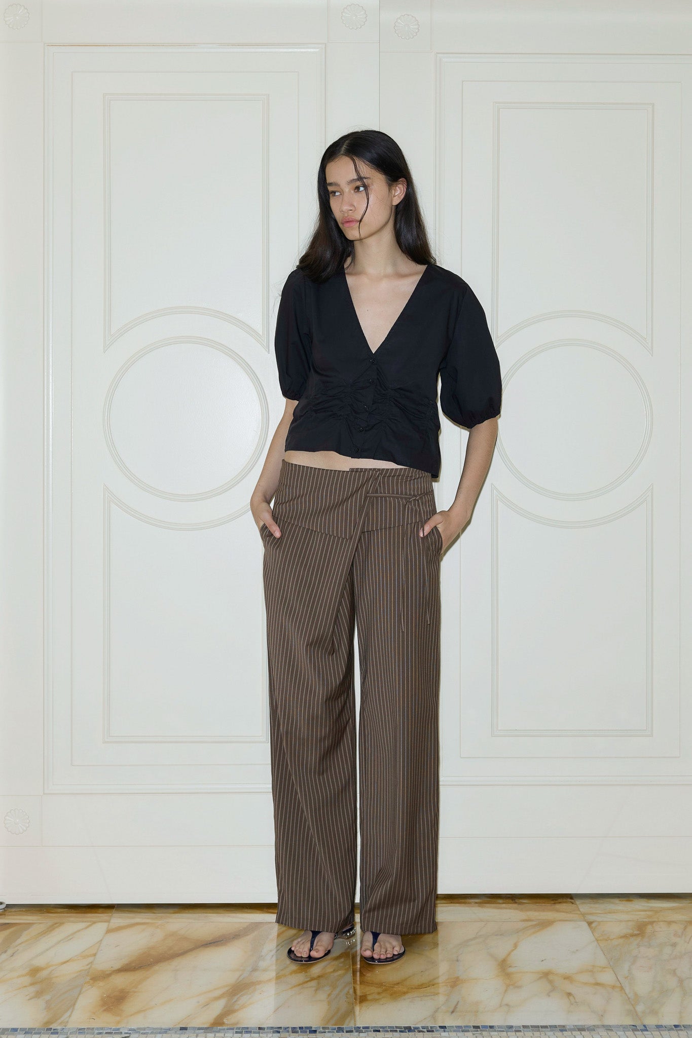 A woman stands indoors, hands in pockets, wearing the 0 Folded Wool Pant - Sable Stripe and a black V-neck short-sleeve top, in front of ornate white double doors.