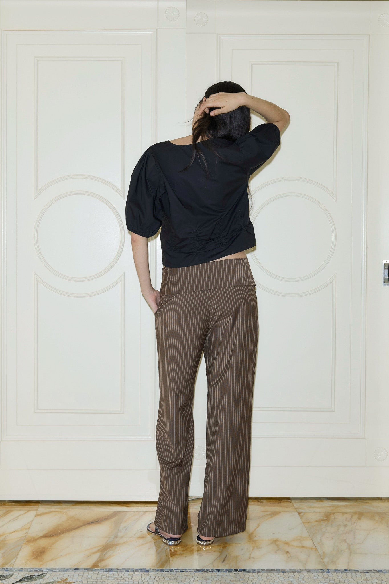 Indoors near white paneled doors, a person in a black top and 0 Folded Wool Pant - Sable Stripe stands with one hand at the back of their head, head slightly turned and face hidden.