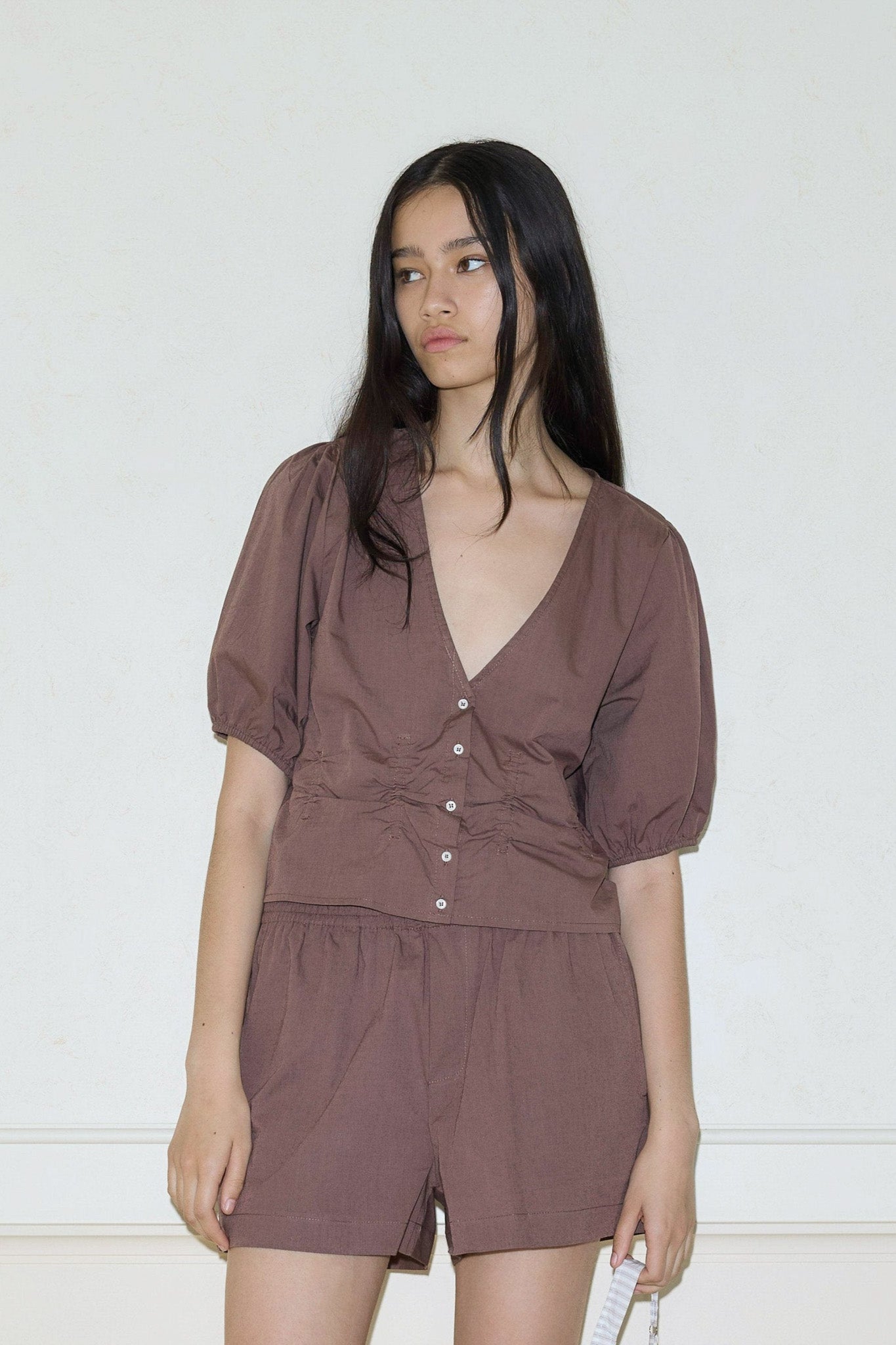 A woman with long dark hair models the Draw Top, a brown button-up blouse with puffed short sleeves, paired with matching brown shorts, standing against a plain light wall and looking slightly to the side.