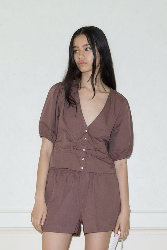A woman with long dark hair models the Draw Top, a brown button-up blouse with puffed short sleeves, paired with matching brown shorts, standing against a plain light wall and looking slightly to the side.