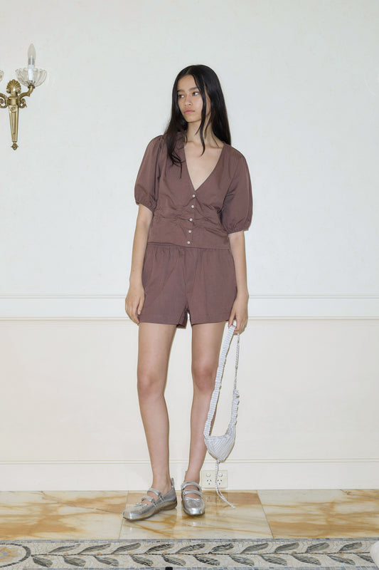 A woman with long dark hair wears the Deiji Studios Organic Cotton Boxer, paired with silver loafers and a silver mini bag. She stands indoors against a light-colored wall with a sconce in the upper left corner.