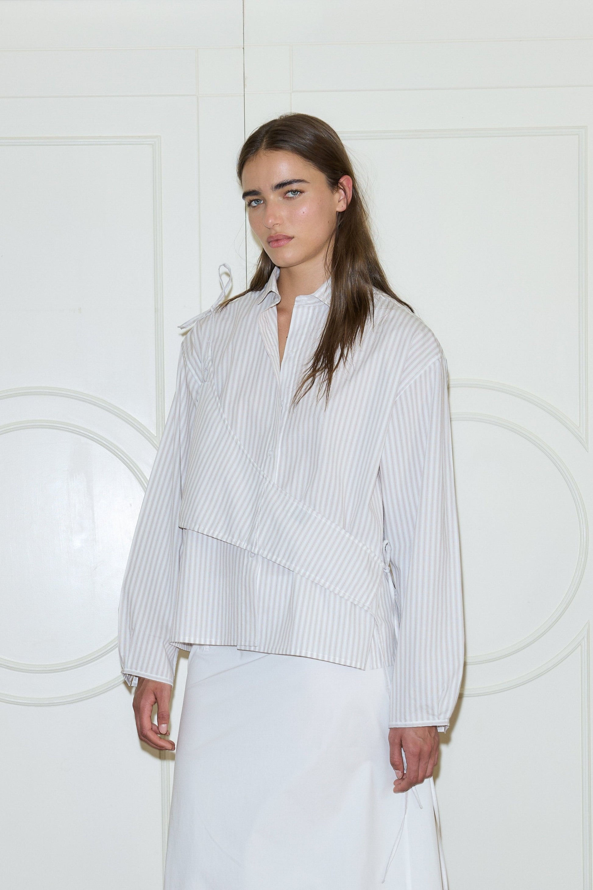 A woman with long brown hair stands before white paneled doors, calmly looking at the camera while wearing the Cotton Sash—a loose, light gray and white striped long-sleeve shirt with a unique layered design—paired with a white skirt.