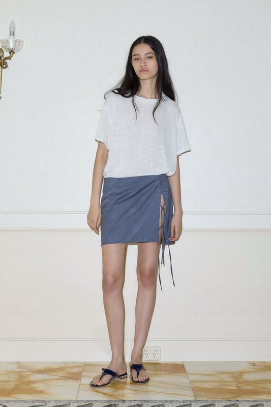 A woman with long dark hair and a neutral expression stands indoors against a light wall, wearing Deiji Studios Two Ties Skirt—a blue pinstriped mini skirt with side strings—paired with a loose white T-shirt and black sandals.