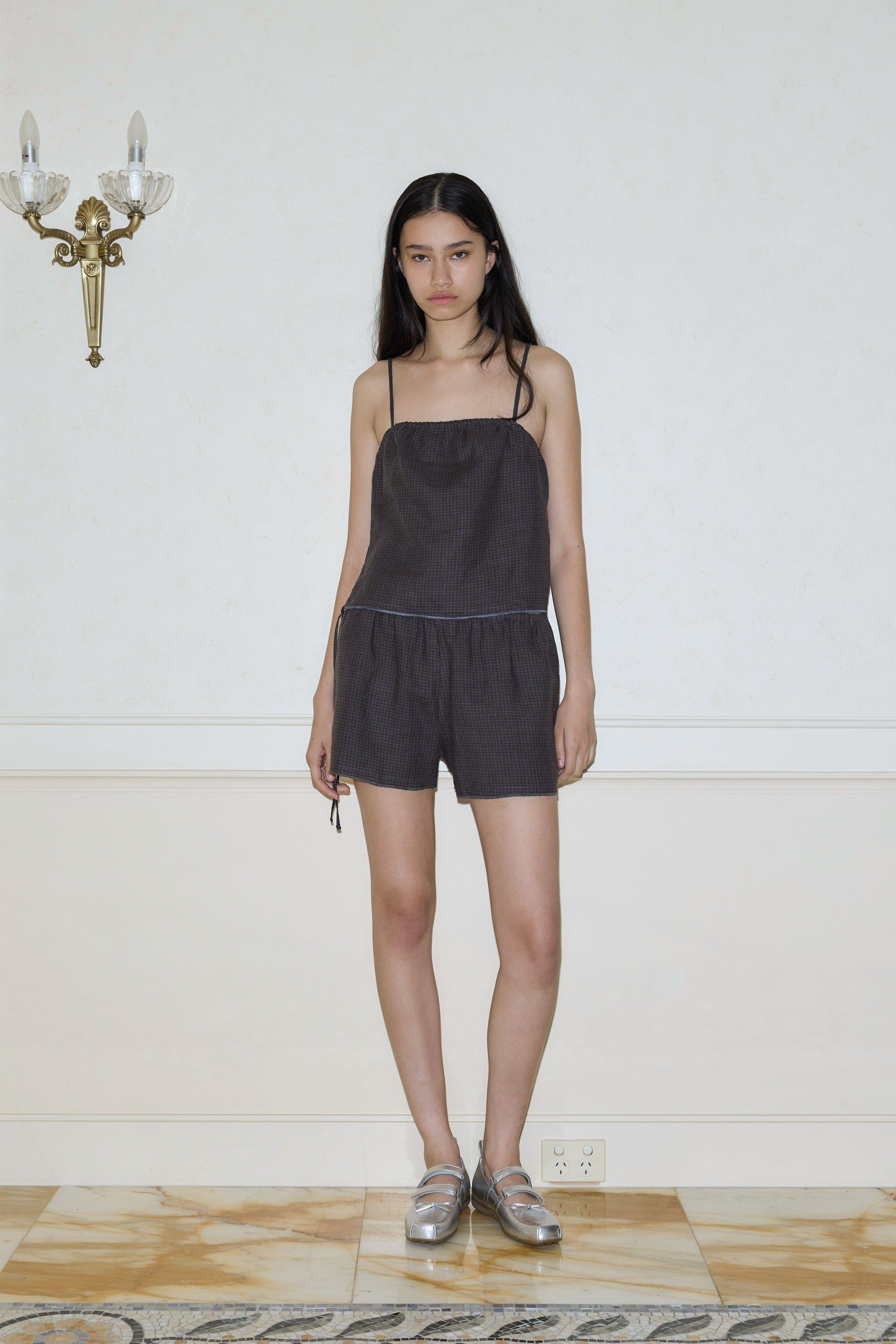 A woman with long dark hair wears the Babylock Linen Set - Slate Check by Deiji Studios, featuring organic linen. She stands indoors against a pale wall with a sconce and marble flooring, completing her look with metallic shoes.