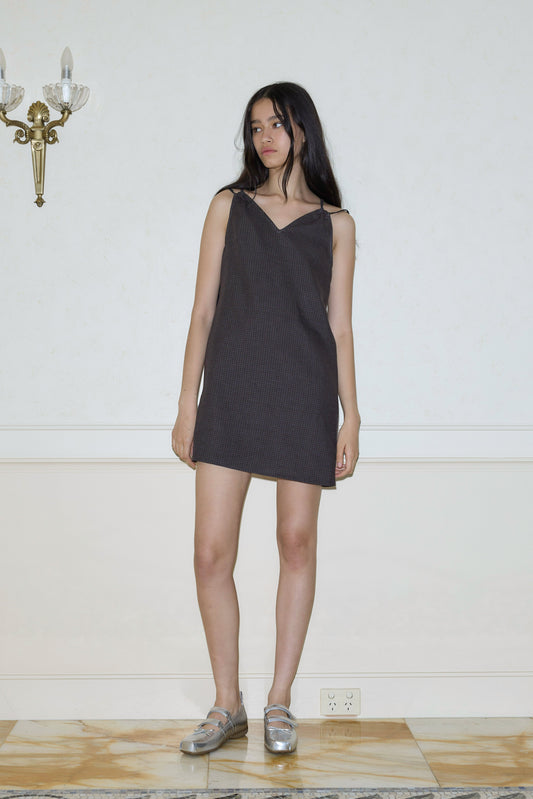 A woman with long dark hair stands indoors against a plain wall, wearing the Looped Dress - Slate Check, which is sleeveless, short, and features a v neckline. She pairs it with silver shoes. A brass light fixture appears above her left shoulder.