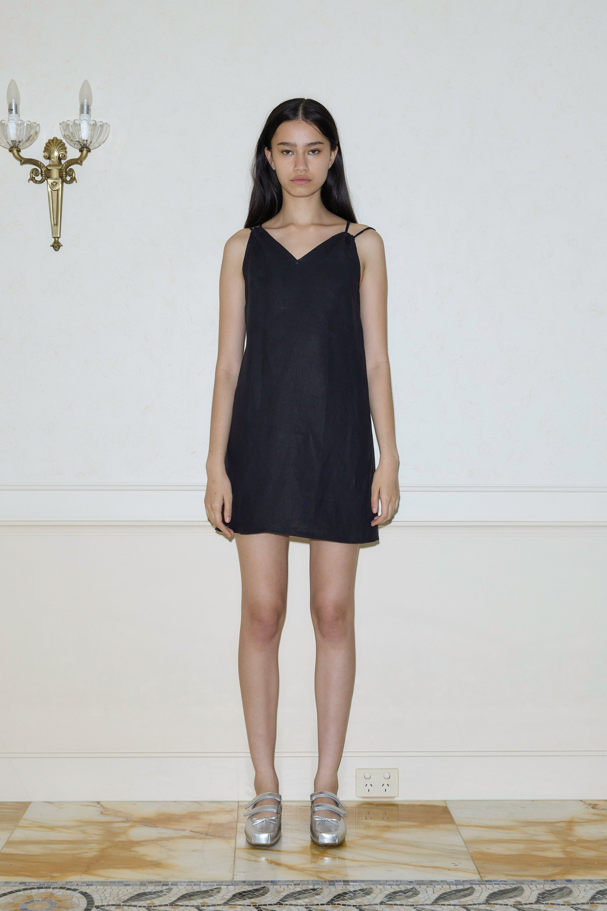 A woman with long dark hair stands indoors against a cream-colored wall, wearing Deiji Studios’ Looped Dress - Black, a sleeveless mini dress, and silver shoes. She gazes neutrally at the camera; a wall sconce appears on the left.
