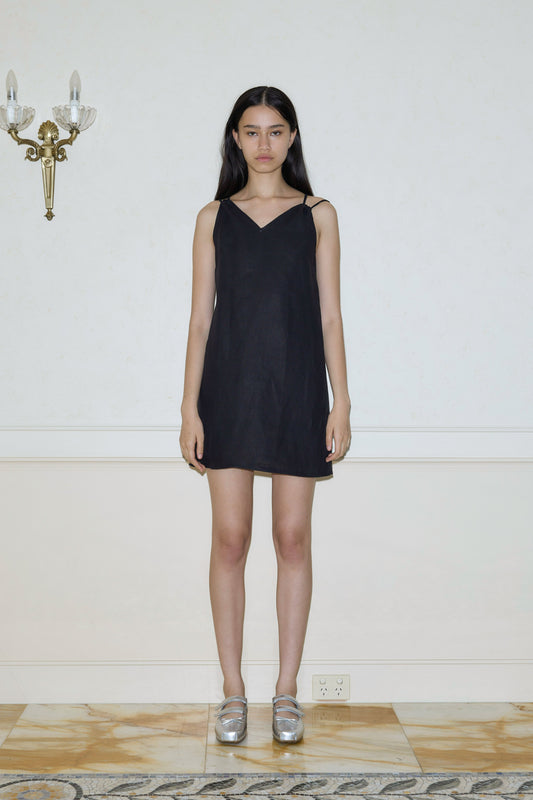 A woman with long dark hair stands indoors against a cream-colored wall, wearing Deiji Studios’ Looped Dress - Black, a sleeveless mini dress, and silver shoes. She gazes neutrally at the camera; a wall sconce appears on the left.