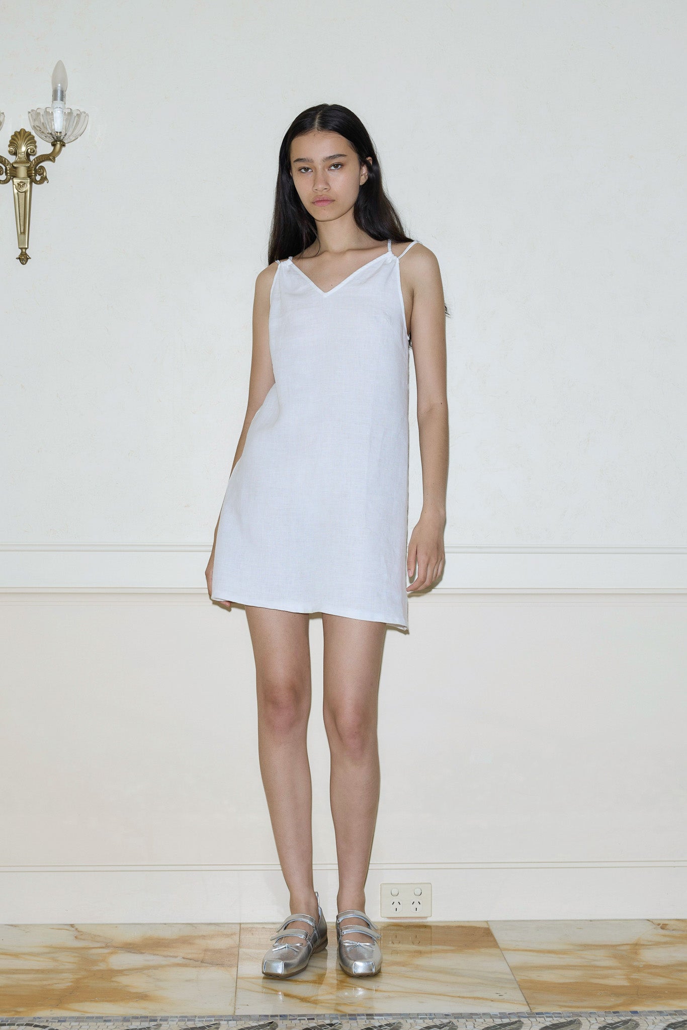 A young woman with long dark hair stands indoors wearing the Looped Dress, a simple sleeveless white piece, paired with silver shoes. She faces forward against a pale wall with a brass sconce and marble floor, her expression neutral.