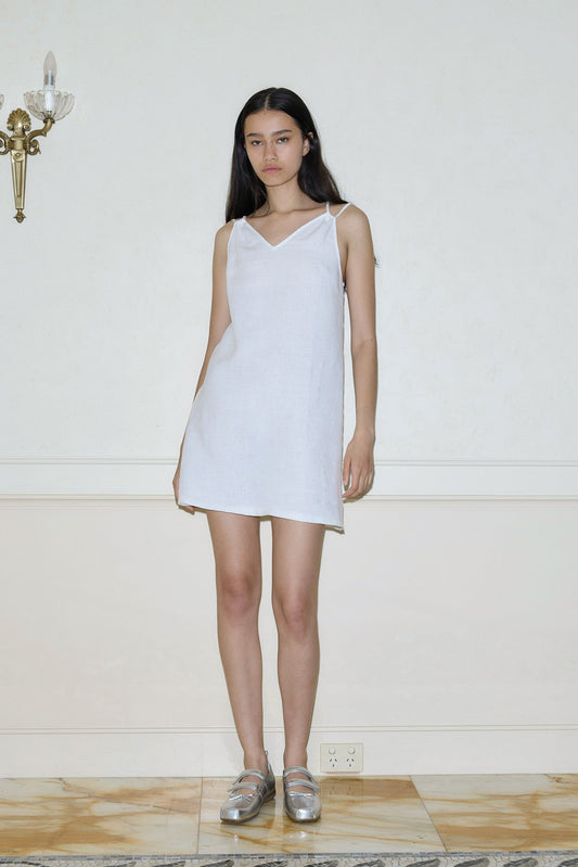 A young woman with long dark hair stands indoors wearing the Looped Dress, a simple sleeveless white piece, paired with silver shoes. She faces forward against a pale wall with a brass sconce and marble floor, her expression neutral.