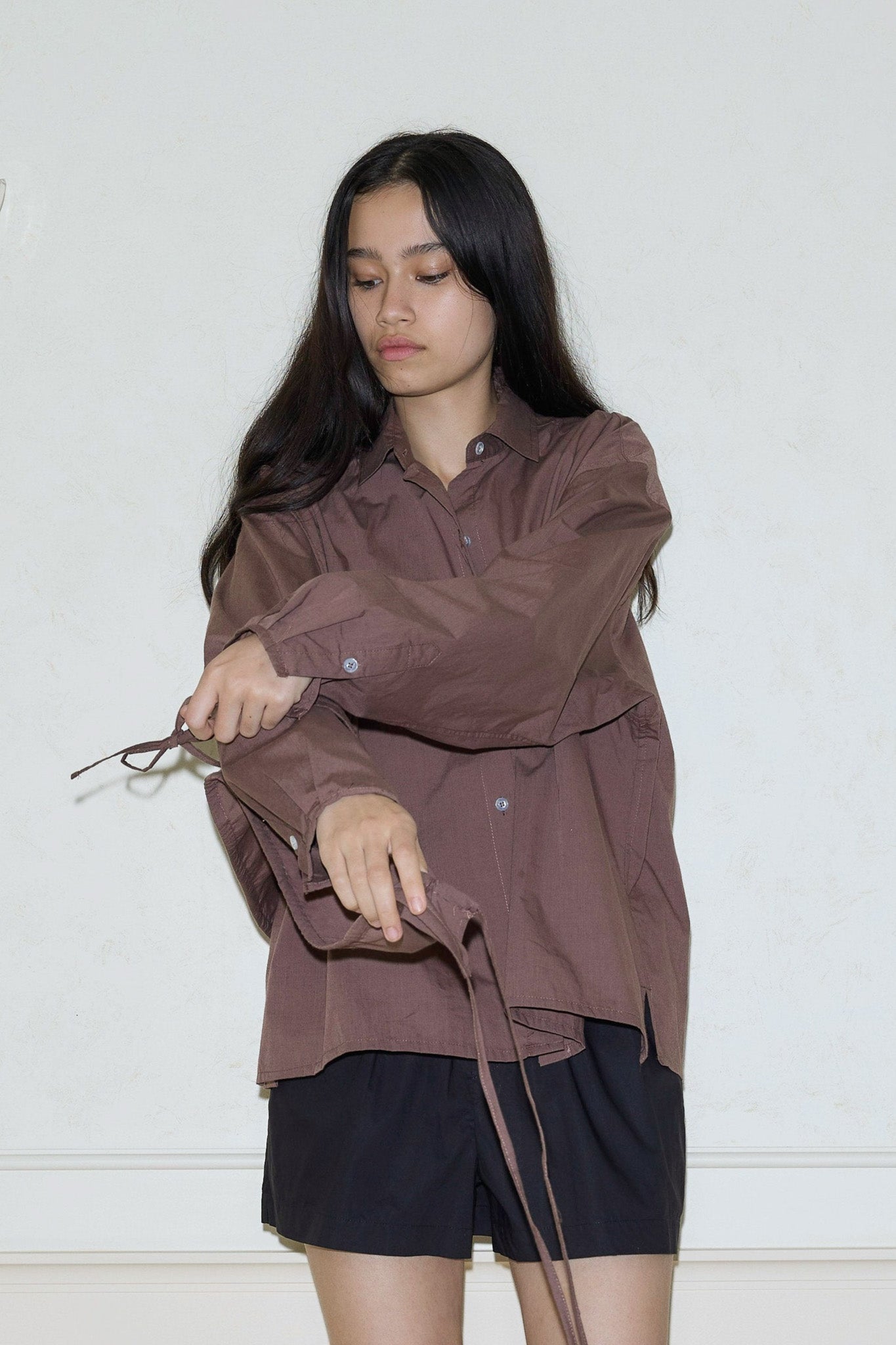A person with long dark hair, wearing a loose brown shirt and black shorts, stands against a light wall, adjusting the Cotton Sash on their shirt and holding a pair of glasses while looking down.