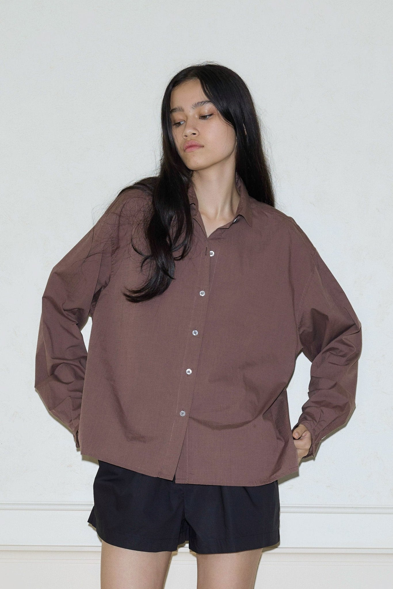A person with long dark hair wears the oversized fit Day Shirt - Cedar from 0 and black shorts, standing against a plain light wall with hands on hips, looking down and to the side.