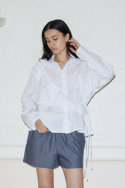 A woman with long dark hair wears a loose white day shirt with drawstring details and dark gray shorts, standing against a plain light-colored wall with a neutral expression.