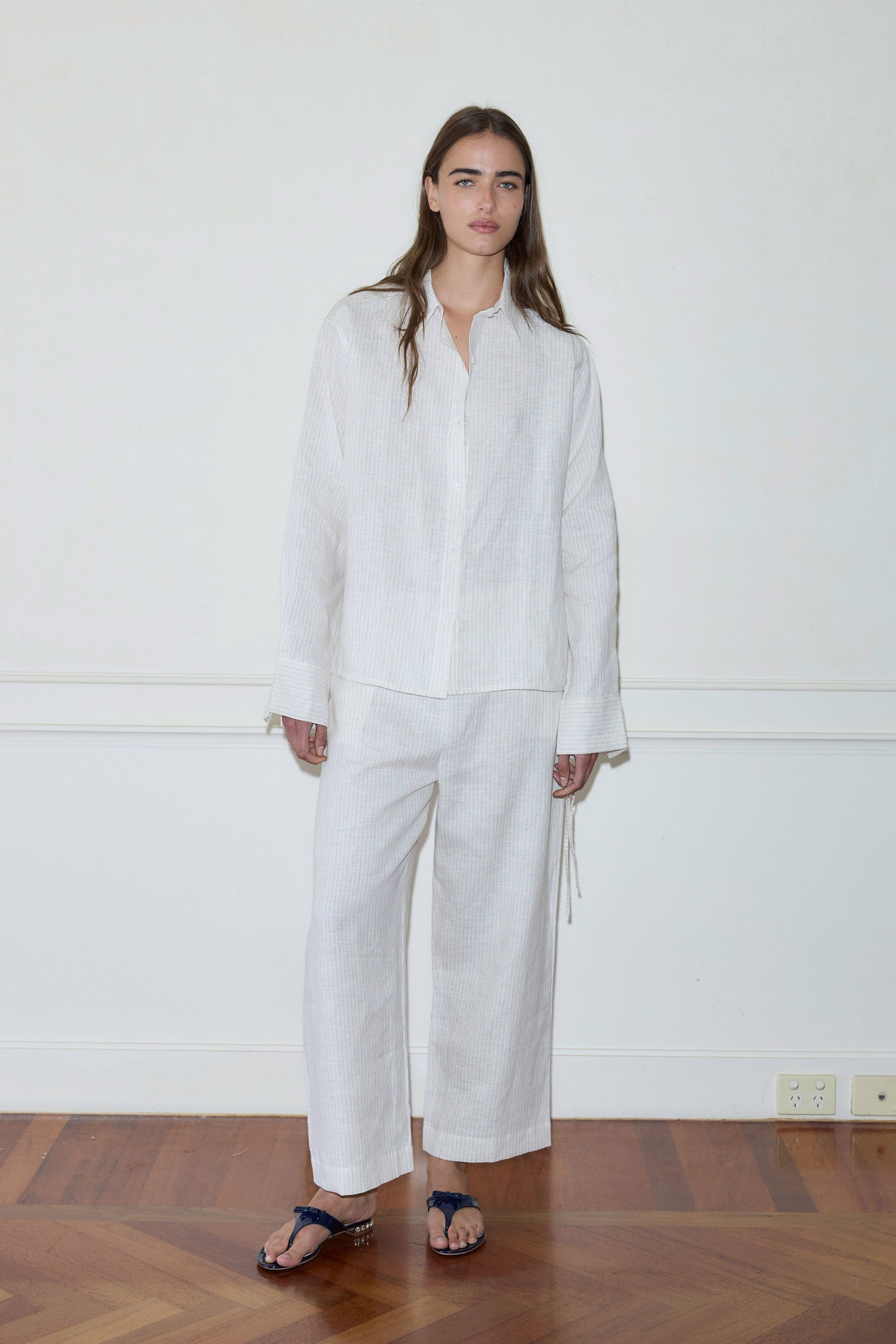 A person stands indoors by a plain white wall, wearing the Tie Split Set—a loose, long-sleeved white shirt and matching wide-legged pants with dark sandals—on a geometric-patterned wooden floor.