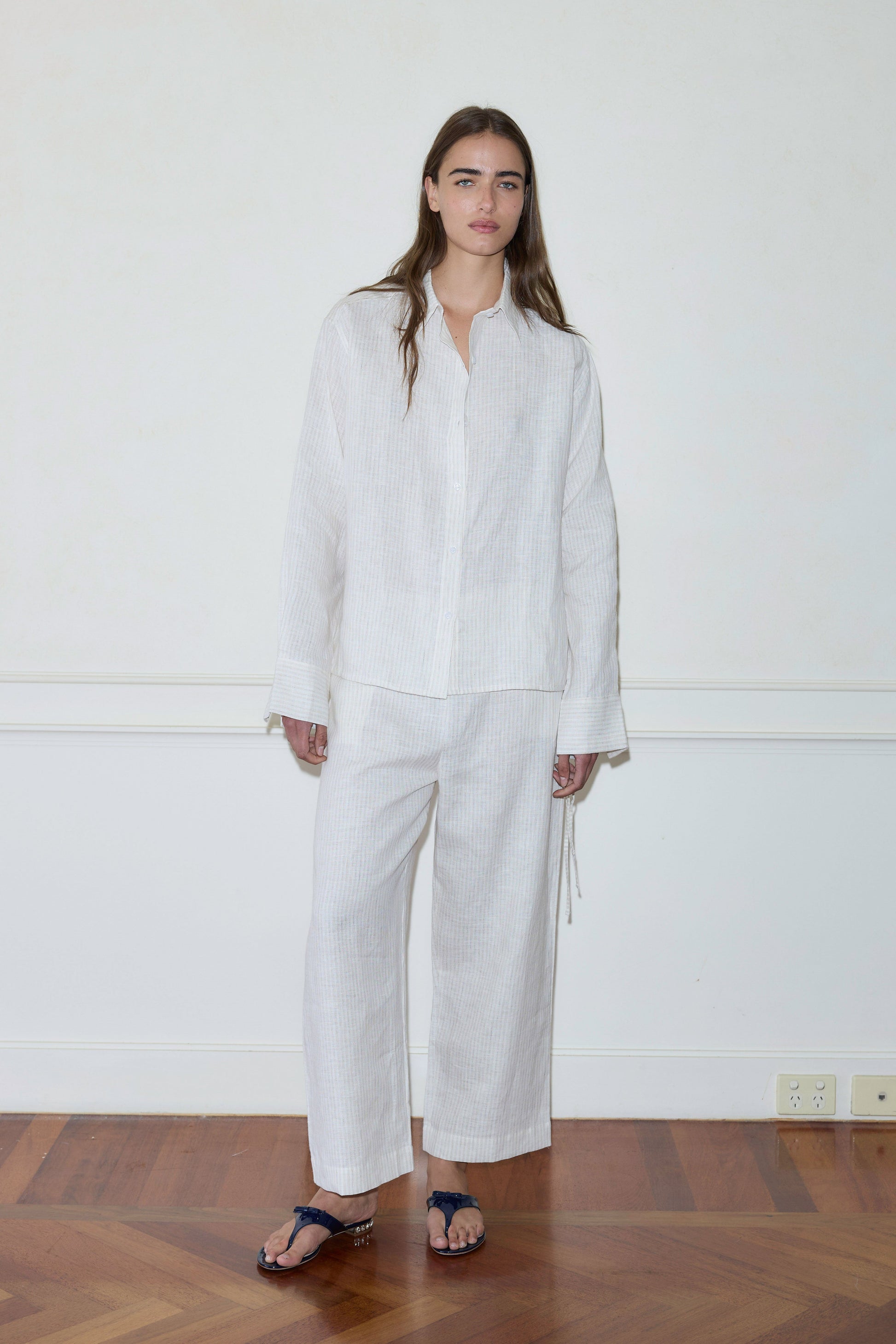 A person stands indoors by a plain white wall, wearing the Tie Split Set—a loose, long-sleeved white shirt and matching wide-legged pants with dark sandals—on a geometric-patterned wooden floor.