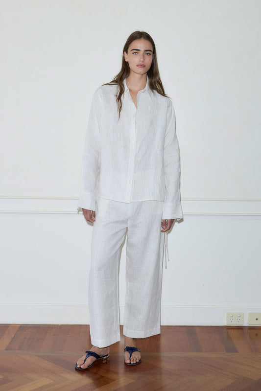 A person stands indoors by a plain white wall, wearing the Tie Split Set—a loose, long-sleeved white shirt and matching wide-legged pants with dark sandals—on a geometric-patterned wooden floor.