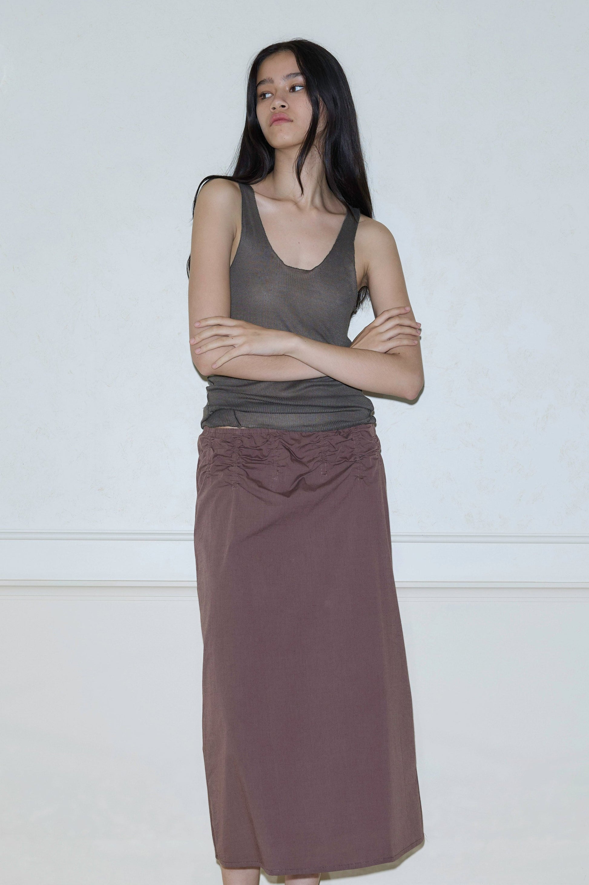 A woman with long dark hair stands against a light wall, wearing a sleeveless olive green top and the Channel Skirt in brown. Her arms are crossed as she looks to the side with a neutral expression.