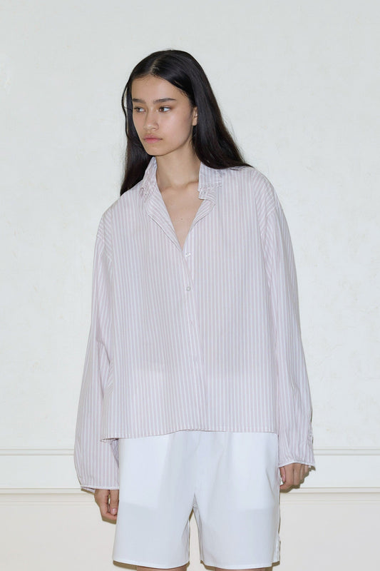 A person with long dark hair wears the day shirt—a loose, light pink and white striped button-up—paired with white knee-length shorts, standing against a plain light-colored wall.