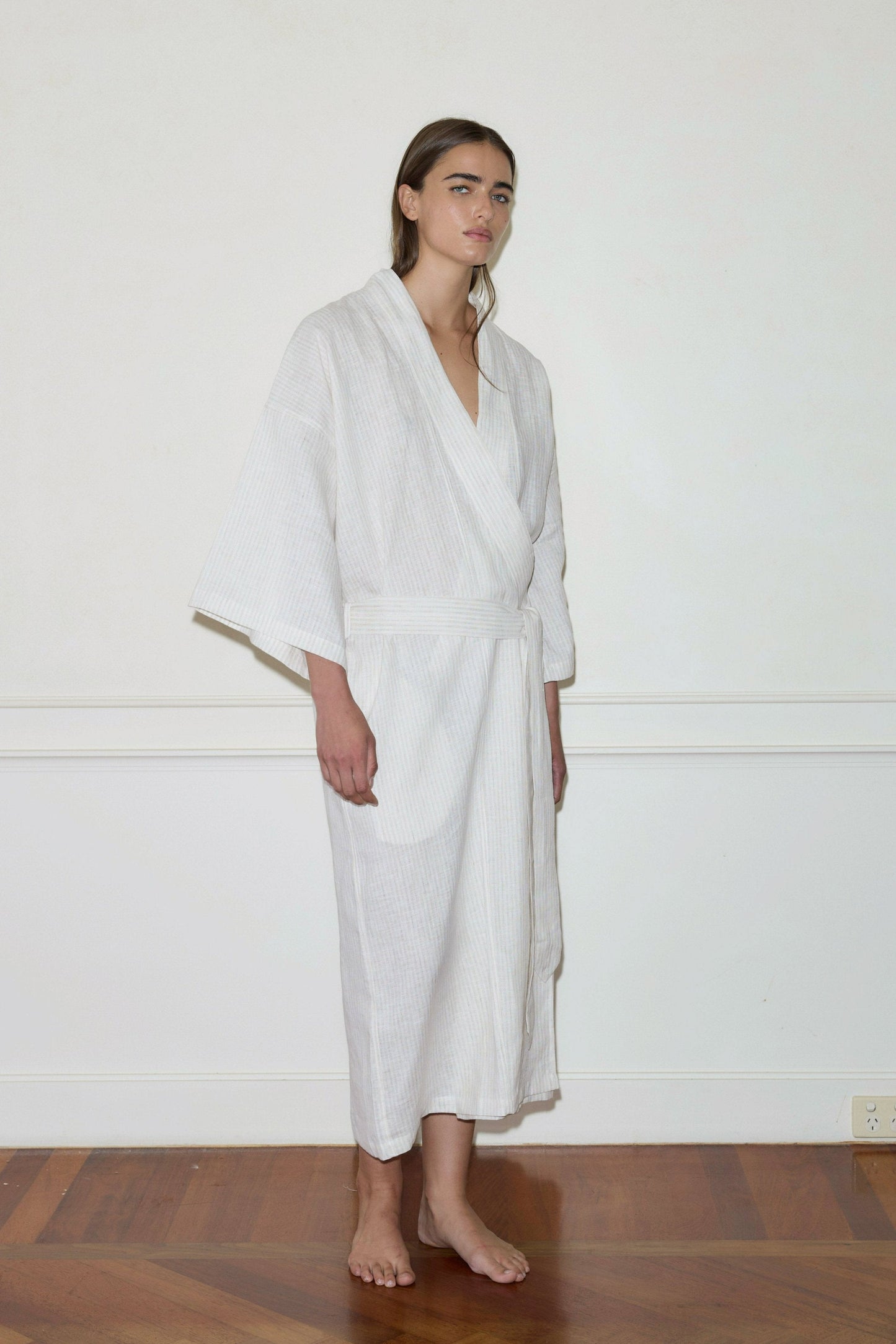 A barefoot person stands indoors on a wooden floor, dressed in the 02 Robe—a long, light-colored garment with loose sleeves—against a plain white wall, gazing slightly to the side.