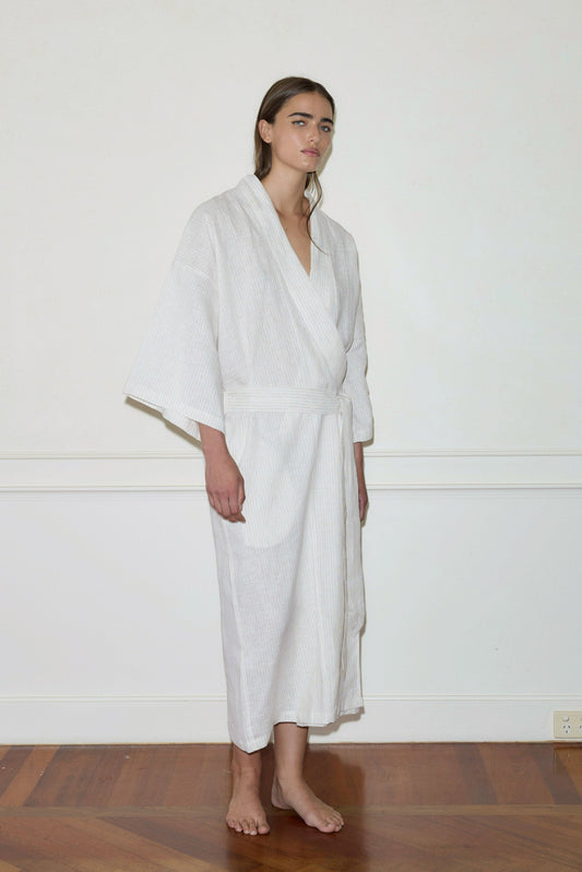 A barefoot person with long brown hair stands indoors against an off-white wall and wooden floor, wearing the 02 Robe—a light, striped, long-sleeve robe featuring large front pockets.