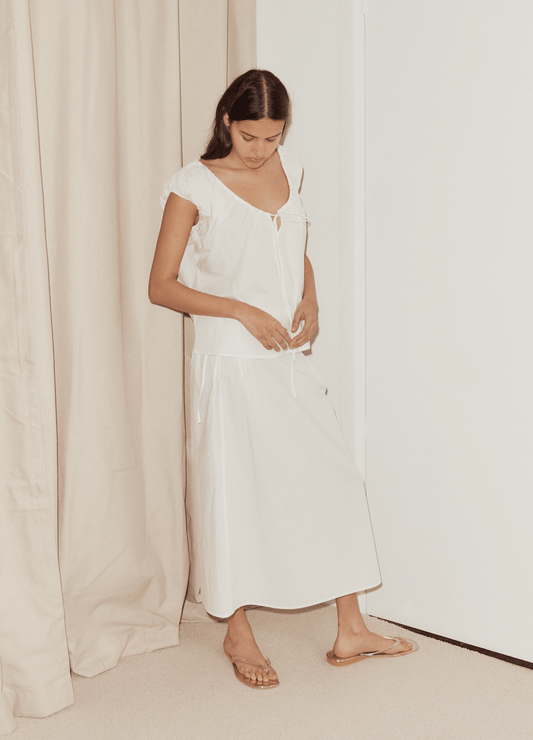 A woman stands indoors near beige curtains and a white wall, wearing a loose white short-sleeve top paired with the Pintuck Skirt and beige sandals. She looks down with her hands near her waist.
