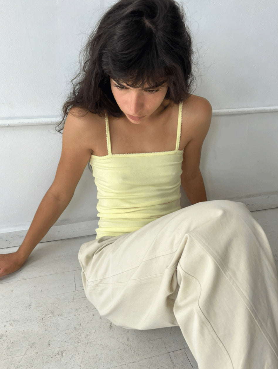 A woman sits on a white studio floor, wearing the Scallop Tank - Lemon by Deiji Studios, a fitted ribbed tank with fine shoulder straps and scallop trim along the straight neckline.