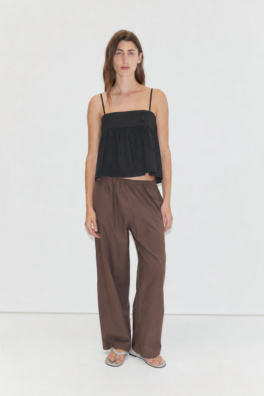 Against a plain white background, a person with long brown hair and a neutral expression models wide brown Ease Trouser pants, paired with a loose black spaghetti-strap top and gray sandals.