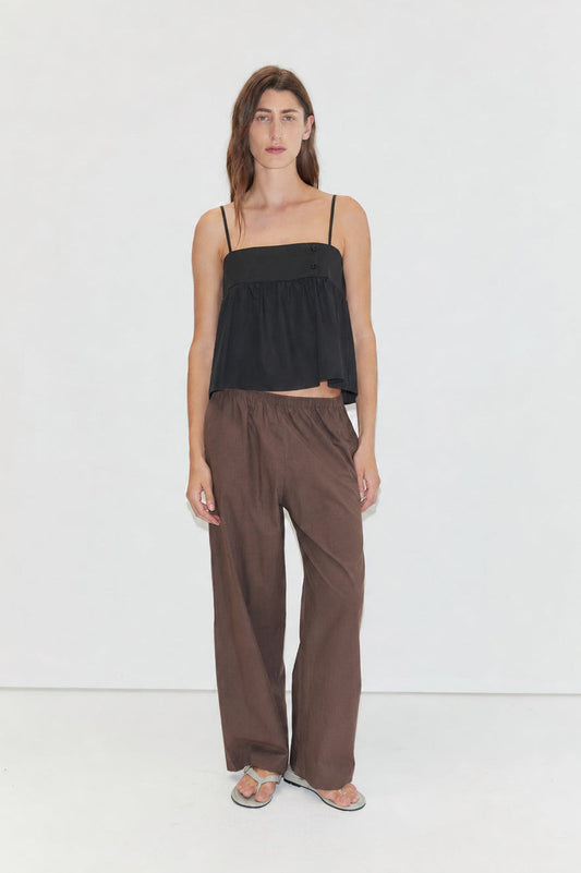 Against a plain white background, a person with long brown hair and a neutral expression models wide brown Ease Trouser pants, paired with a loose black spaghetti-strap top and gray sandals.