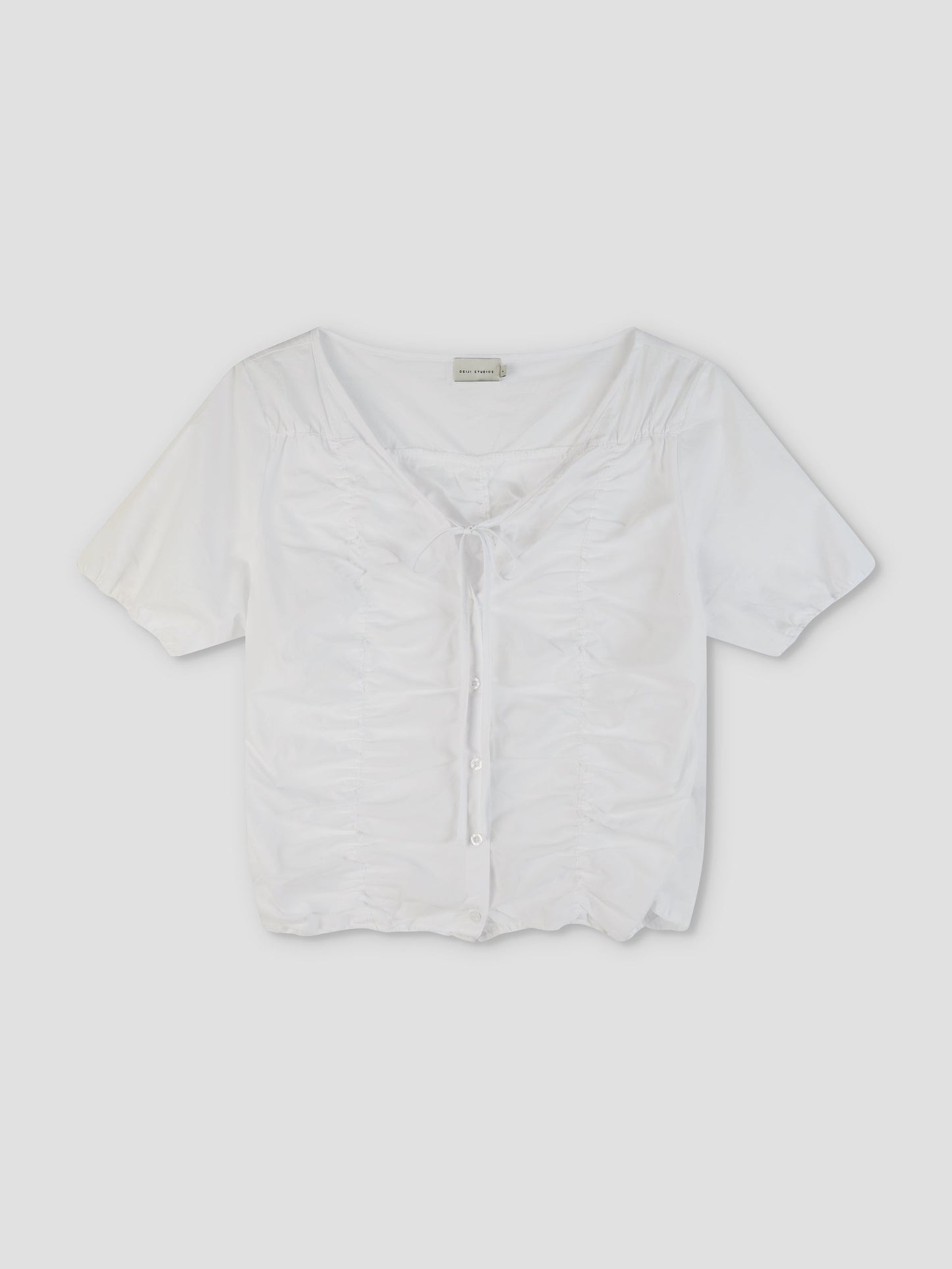 The Crinkle Top - White is a short-sleeved, white button-up top featuring a ruched front, tie detail at the neckline, and a slightly cropped hem, displayed against a plain light background.