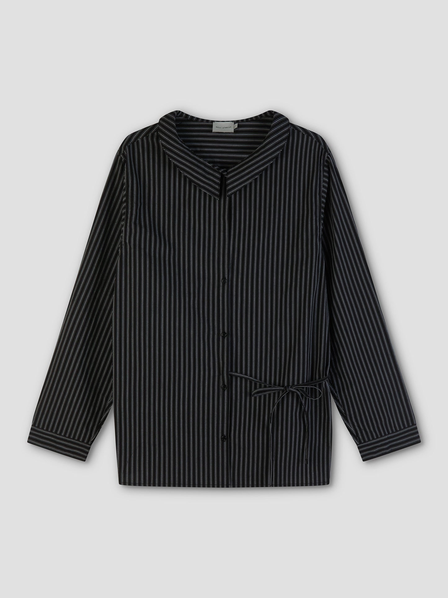 The Left Shirt - Mono Stripe is a black, long-sleeve lightweight shirt with vertical gray pinstripes, wide collar, front buttons, and a small adjustable side tie at the waist, shown on a plain light gray background.