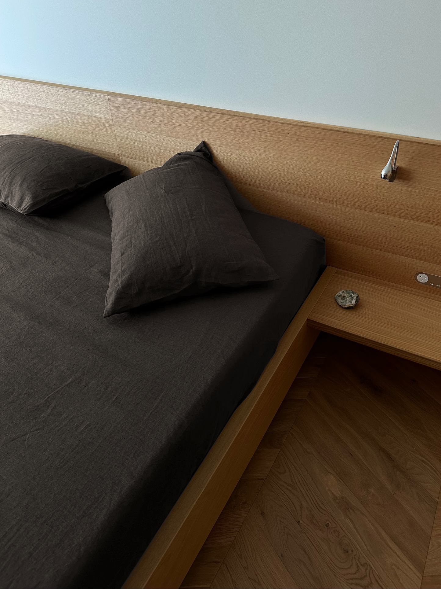 A neatly made bed with Pillow slip set - Bark in deep brown lies against a light wooden headboard, with a small attached side table holding a stone and electronic button—an arrangement inspired by Deiji Studios’ serene style.