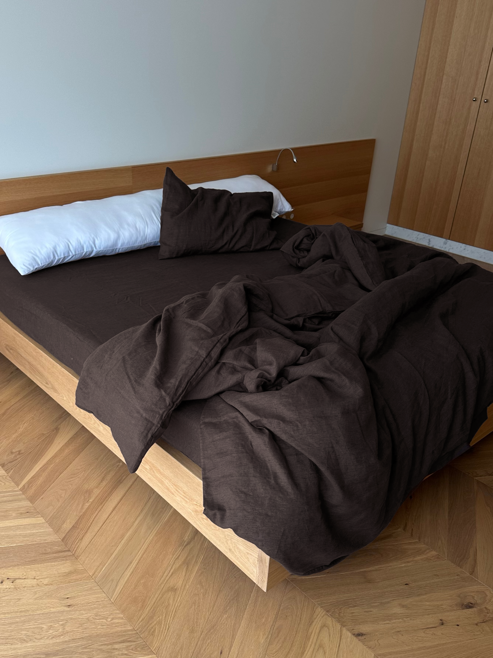 A wooden bed with a dark brown duvet features Deiji Studios’ Pillow slip set - Bark, an unarranged brown pillow, and a long white pillow against the wooden headboard. The unmade bed has blankets partially hanging off the side.