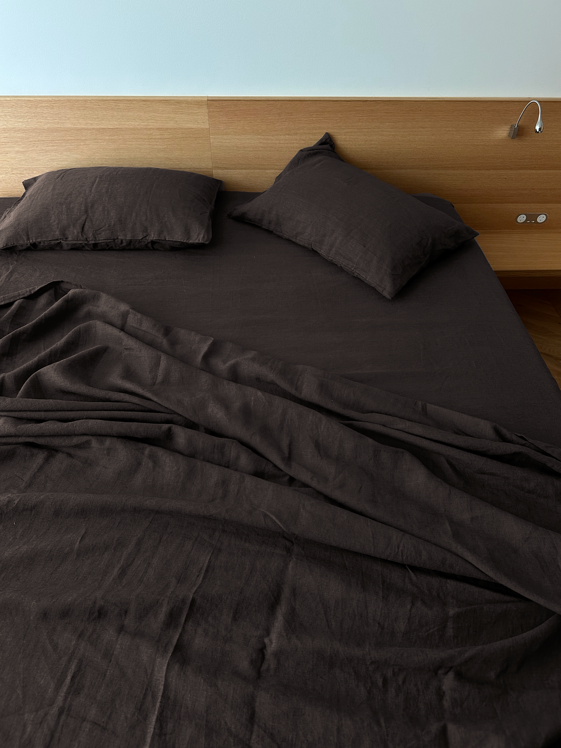 An unmade modern bed features Deiji Studios' Pillow slip set - Bark, with dark brown sheets, hypo-allergenic bedding, and two matching linen pillow slips. The wooden headboard includes a built-in reading light and an electrical outlet on the right.