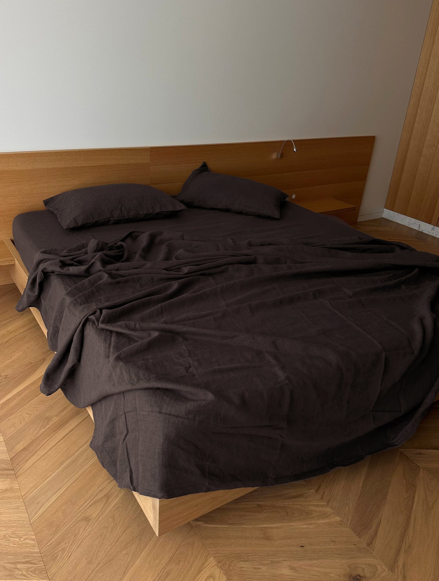 A minimalist bedroom features a modern bed with Deiji Studios’ hypo-allergenic Pillow slip set - Bark, brown sheets, a wooden headboard, light wood flooring, and plain white walls. The linen bedding looks slightly rumpled.
