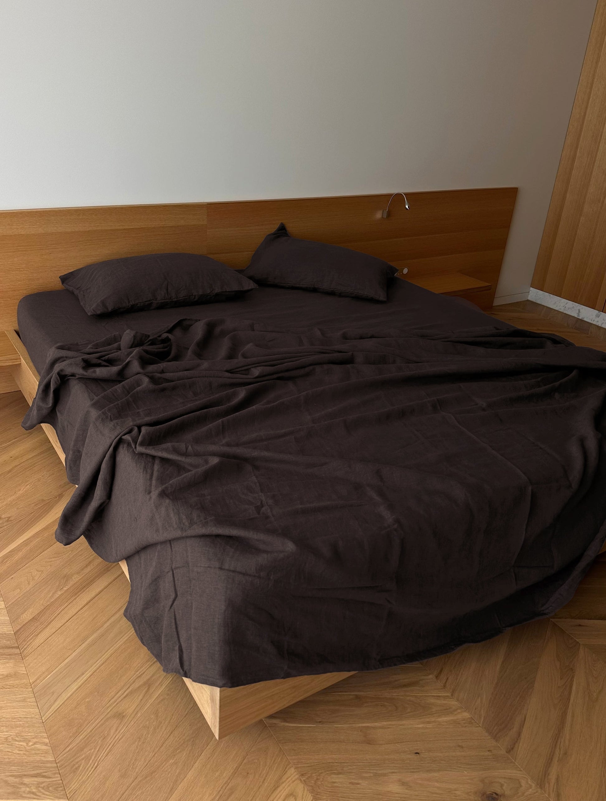 A minimalist bedroom features a modern bed with Deiji Studios’ hypo-allergenic Pillow slip set - Bark, brown sheets, a wooden headboard, light wood flooring, and plain white walls. The linen bedding looks slightly rumpled.