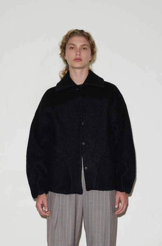 A person with curly hair wears the Boiled Wool Coat, a loose-fitting black button-up jacket, paired with gray pinstriped trousers against a plain white background.