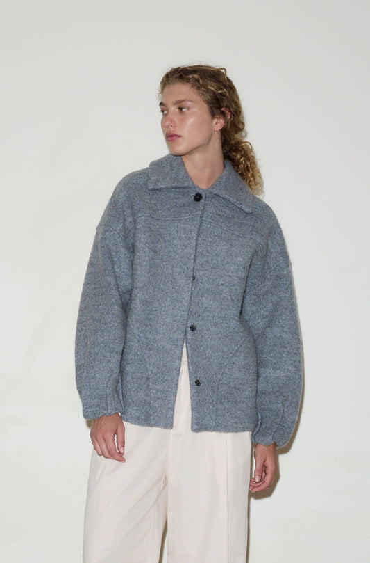 A person with curly hair wears the Boiled Wool Coat, a loose, light gray button-up jacket with large sleeves, paired with cream-colored pants, standing against a plain white background.