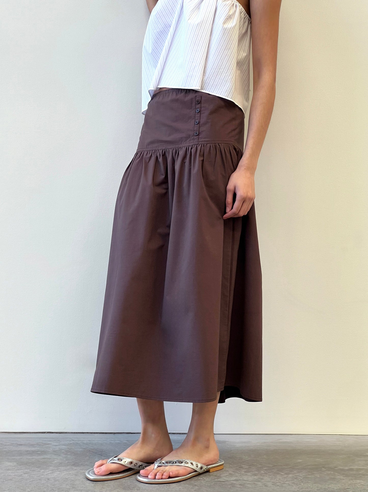 A person wearing a white sleeveless top, the Button Skirt - Chocolate (a high-waisted organic cotton skirt with brown tones), and beige flip-flops stands against a plain light-colored wall.