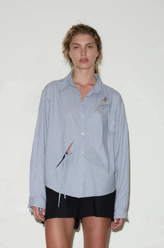 A person with curly hair wears the twin bow shirt, a loose light blue pinstriped button-up with cut-out details, and black shorts while standing against a plain white background.