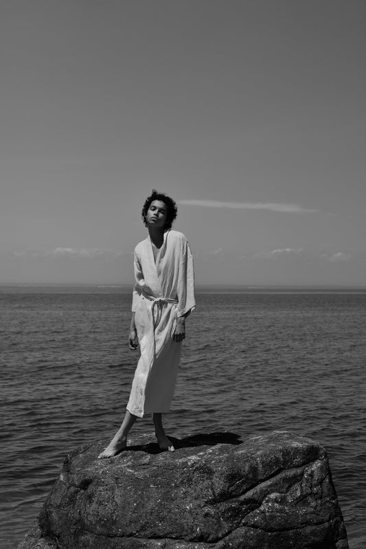 Female model standing on rock wearing the 02 robe - oatmeal by Deiji Studios against ocean horizon