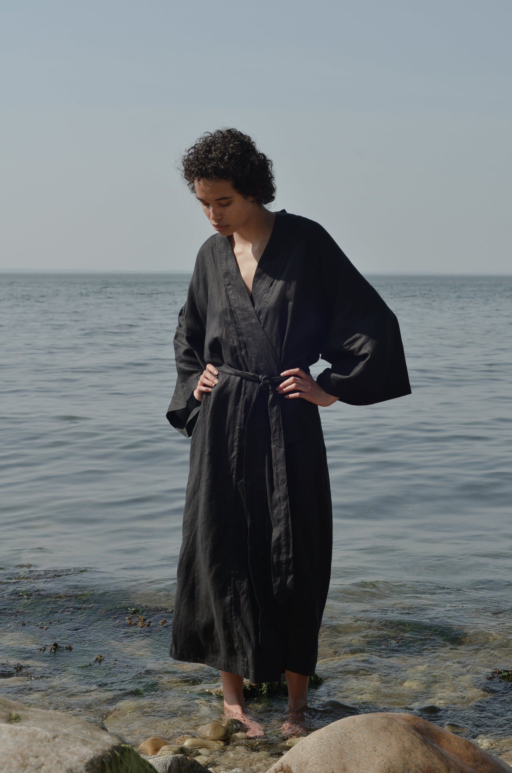 A person with short curly hair stands on a rocky shore, wearing the 02 Robe—a long black robe with wide sleeves—looking down with hands on hips, as the calm sea and a clear sky fill the background.