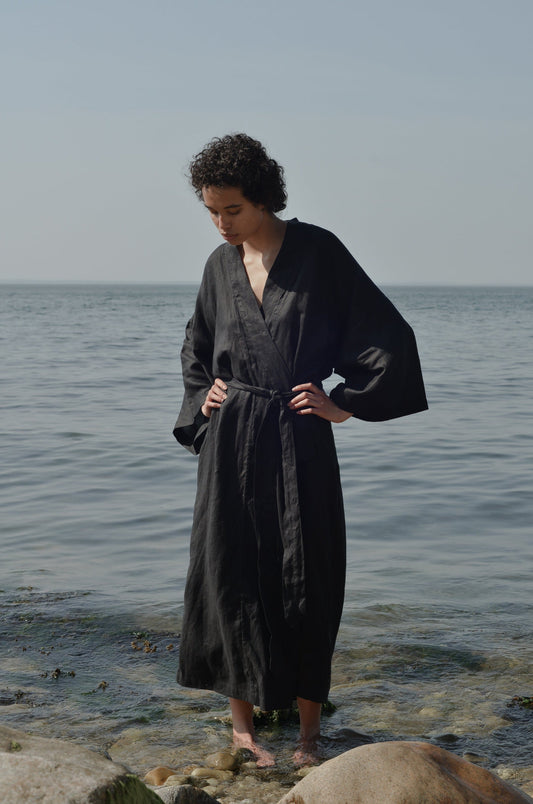 Wearing the 02 Robe, a person with short curly hair stands on a rocky shore, hands on hips, looking down at the calm sea and sky in the background.