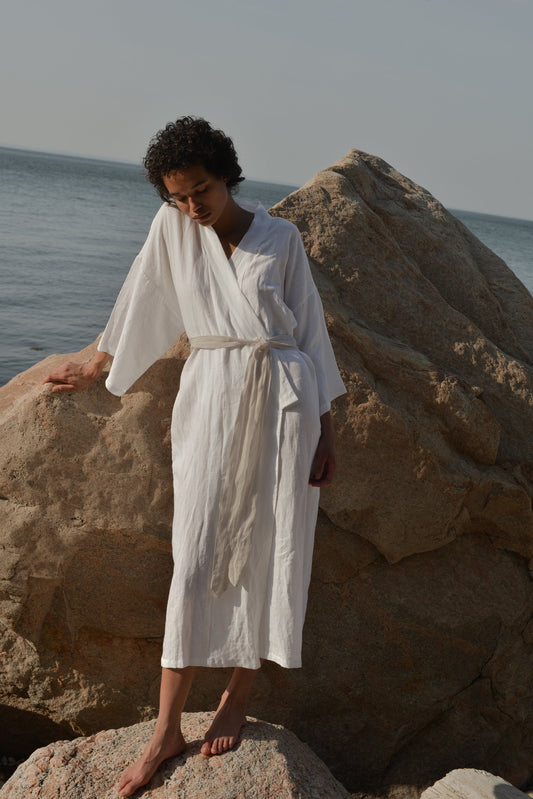 A person wearing the 02 Robe stands barefoot on rocks by the sea, with large boulders and calm water under a clear sky in the background.