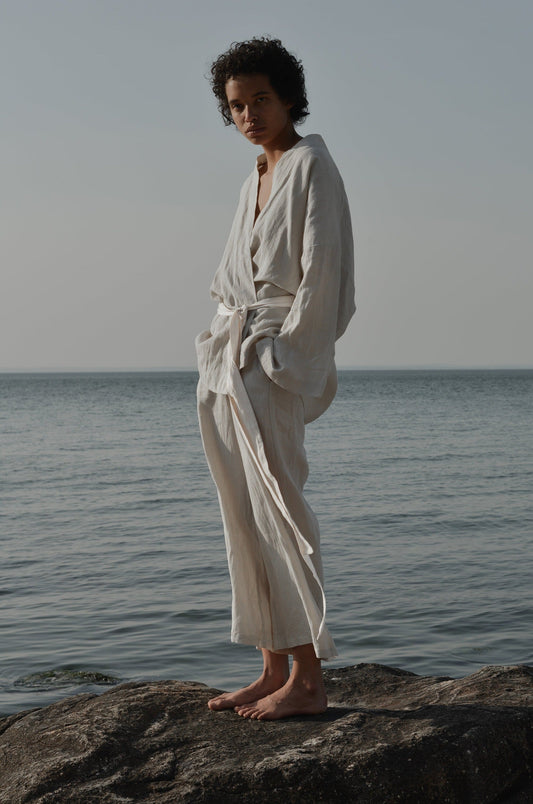 Barefoot on a seaside rock, a person wears the 01 Set—loose, light-hued long-sleeve robe and pants—framed by calm water and an overcast sky for a serene, minimalist look.