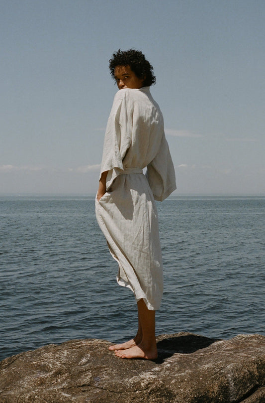 Female model standing on rock wearing the 02 robe - oatmeal by Deiji Studios against ocean horizon